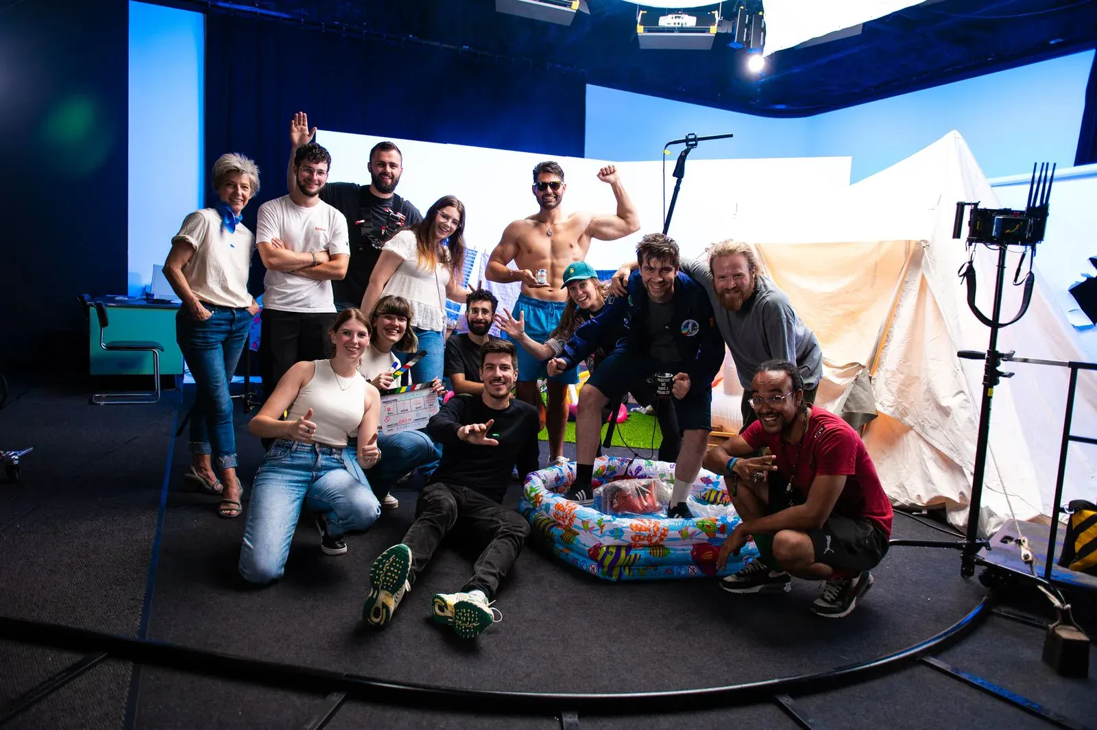 Het GROM-team poseert op een filmset, rond een opblaasbaar zwembad en een tent